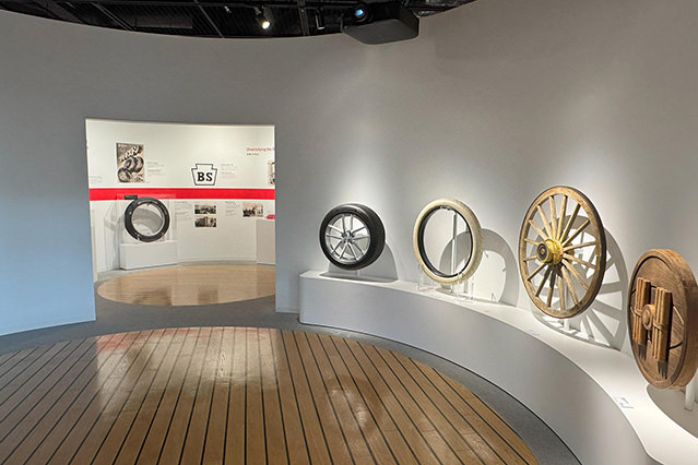 Curved museum display showing historical Bridgestone wheels and tire designs mounted on a white wall, with wooden flooring in the foreground.