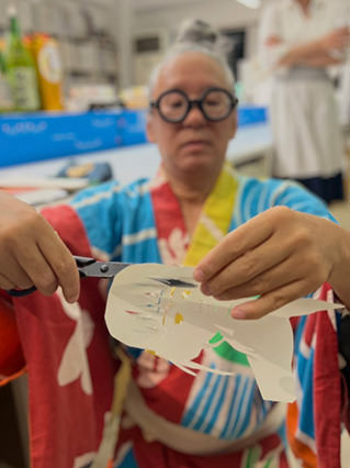 Artist wearing round glasses and a colorful yukata, carefully cutting paper with scissors to create a kirie artwork.
